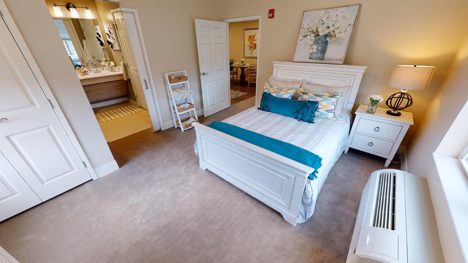 A bright and cozy bedroom in a senior living facility featuring a white wooden bed with colorful pillows and a teal throw blanket. Next to the bed is a white nightstand with a lamp and a vase of flowers. A window with natural light is on the right side, and an air conditioning unit is below it. The room has beige walls and carpeted floors. An open door leads to a bathroom with a vanity and mirror, and another door opens to a dining area with a table and chairs.