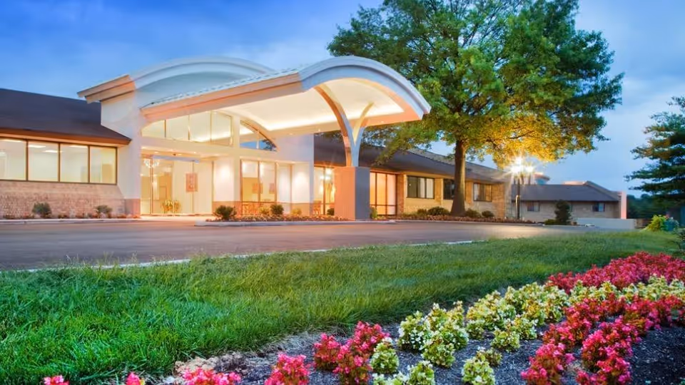 Front entrance of a senior living facility with a covered porte-cochère, illuminated windows, lawn and flowerbeds.