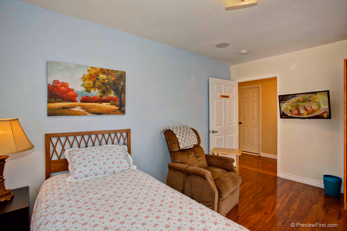 A bedroom with a single bed covered in a white and pink floral bedspread, a wooden headboard, a brown recliner chair with a patterned blanket draped over the back, a small wooden side table, a lamp on a black nightstand, a wall-mounted TV showing a food program, and a painting of a landscape with trees on the light blue wall.