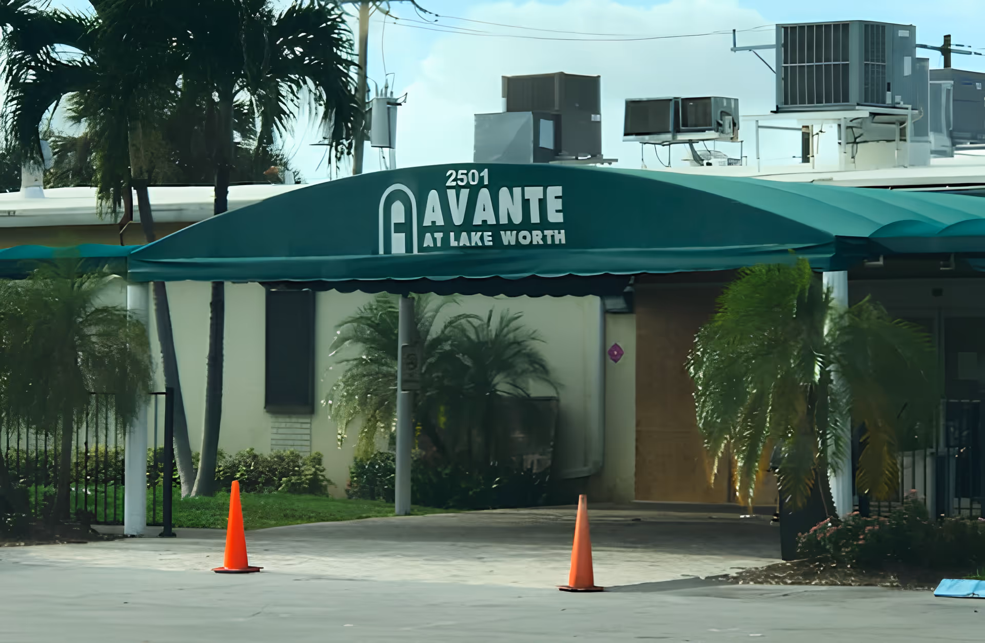 Entrance canopy of Avante at Lake Worth Rehabilitation and Skilled Nursing facility with green awning displaying the facility name and address number 2501, surrounded by palm trees and orange traffic cones on the driveway.