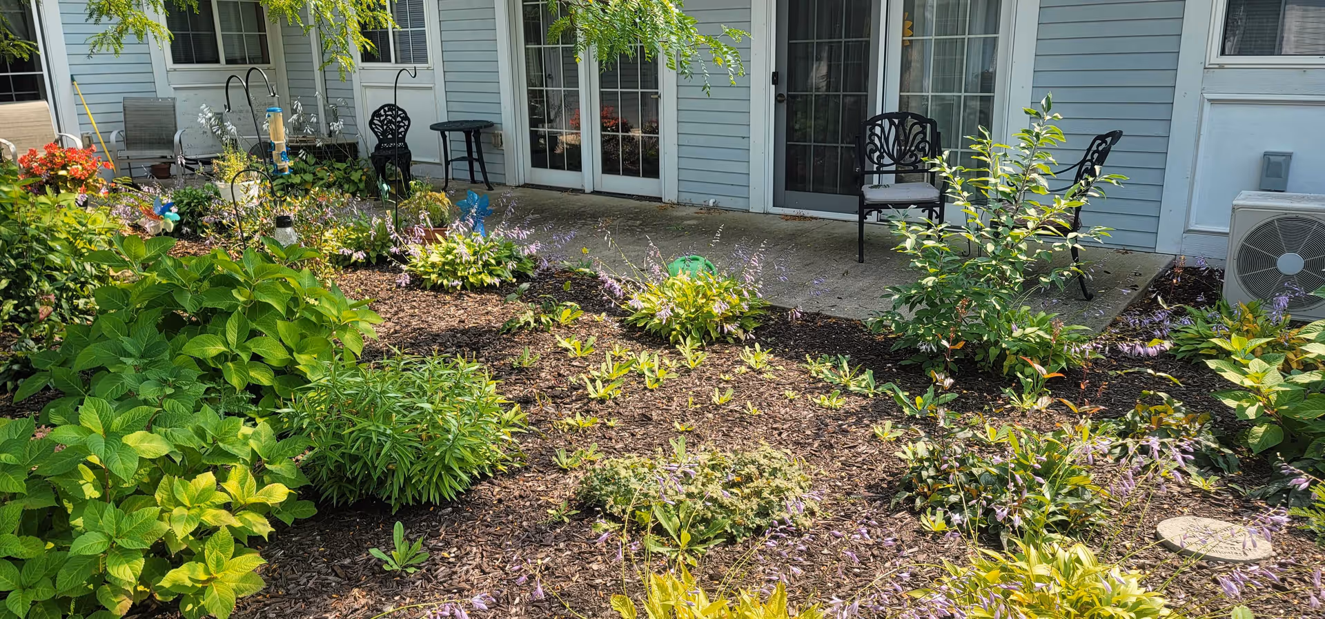 A garden area with various green plants and flowers in front of a building with light blue siding. There are two black metal chairs and a small black table on a concrete patio near glass doors. An air conditioning unit is visible on the right side of the image.
