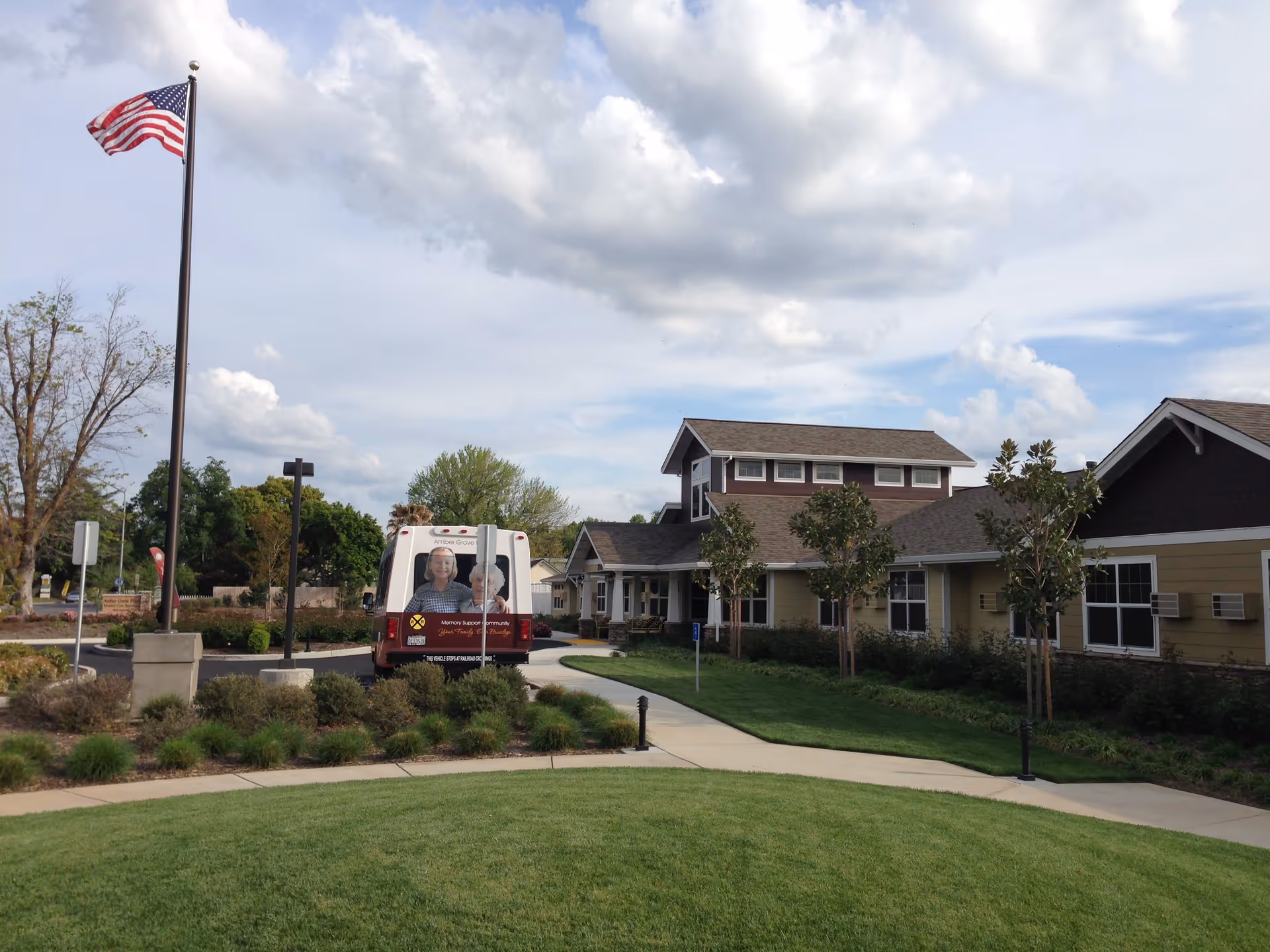 Exterior view of Amber Grove Place Memory Care facility with a well-maintained lawn, a paved walkway, several small trees, and a building with beige siding and a brown roof. An American flag is flying on a flagpole to the left, and a van with images of elderly people and the facility's name is parked near the building.