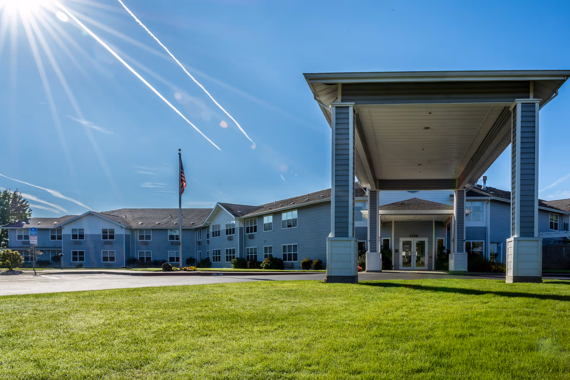 Front entrance of a two-story assisted living building with a covered porte-cochere, an American flag, and a manicured lawn under a sunny sky.