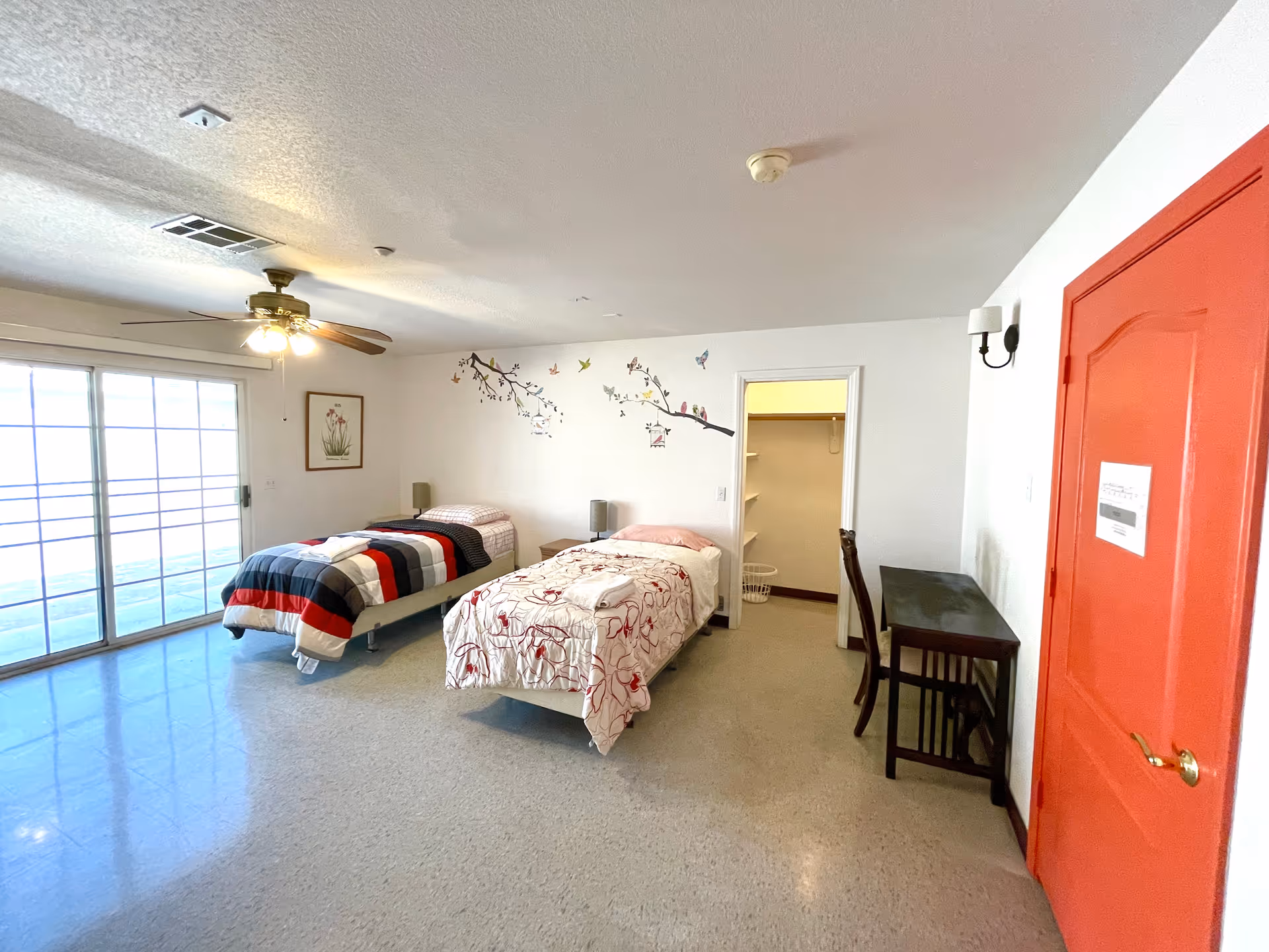 Bright assisted-living bedroom with two twin beds, sliding glass doors, a ceiling fan and a red entry door.