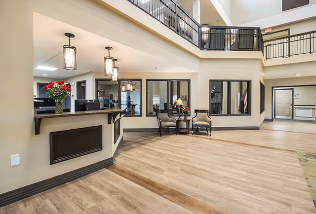 Spacious and well-lit reception area in a senior living facility with a wooden floor, a front desk adorned with a vase of red roses, two chairs and a small table with a lamp and flowers, large windows, and a second-floor balcony overlooking the area.