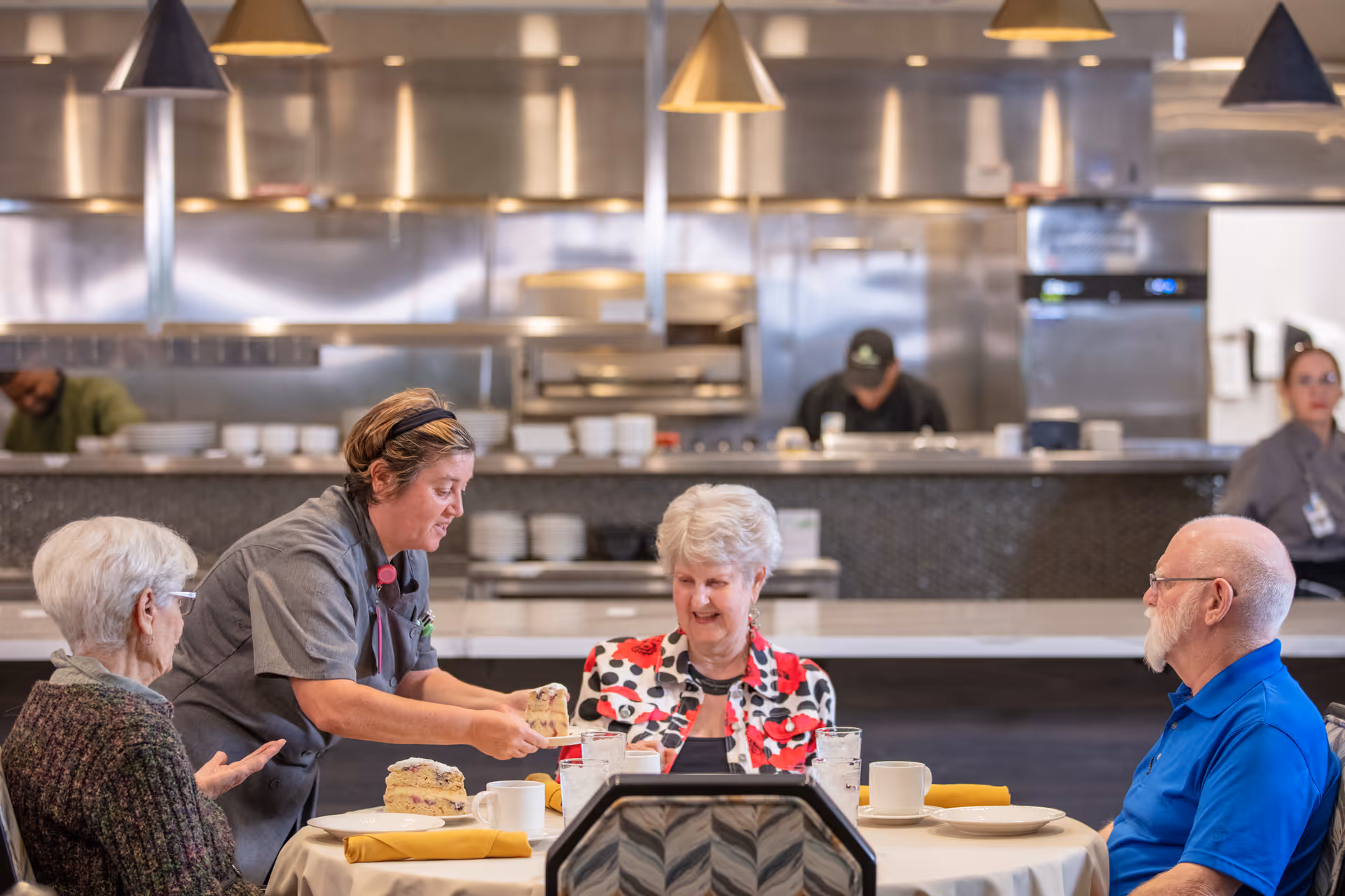 A dining area in a senior living facility where a staff member is serving a slice of cake to three elderly individuals seated around a table set with plates, cups, and yellow napkins. The background shows a kitchen area with stainless steel appliances and other staff members working.