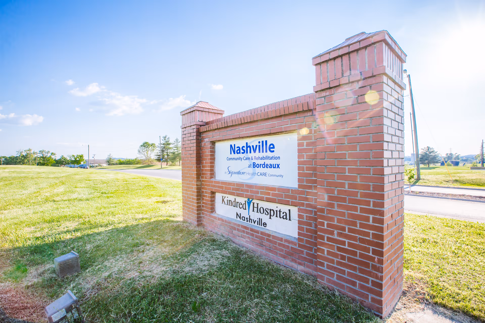 A brick signpost on a grassy area beside a road displaying the names Nashville Community Care & Rehabilitation at Bordeaux and Kindred Hospital Nashville under a clear blue sky.
