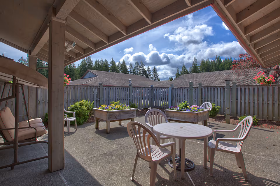 Outdoor patio area with a round white plastic table surrounded by four white plastic chairs. There are two raised garden beds with colorful flowers, a wooden swing with cushions, and a wooden fence enclosing the space. Trees and a partly cloudy sky are visible in the background.