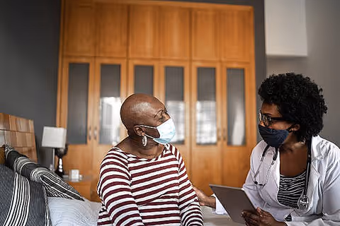 An elderly woman wearing a striped shirt and a face mask sits on a bed while talking to a healthcare professional who is also wearing a face mask and holding a tablet. They are in a room with wooden cabinets and a bedside lamp.