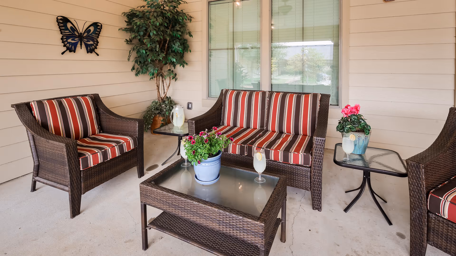 A cozy outdoor seating area with wicker furniture featuring red and white striped cushions. There is a loveseat, two armchairs, a glass-top coffee table with a potted plant, and a side table with a pink flowering plant. A decorative butterfly wall art and a tall potted plant are against the beige siding wall, with windows in the background.