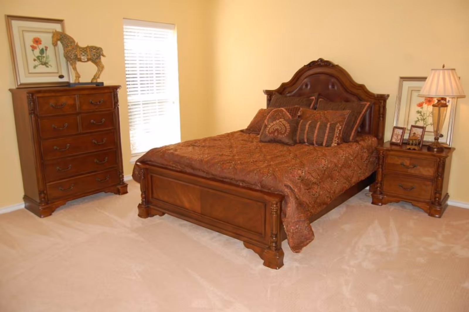A bedroom with a wooden bed frame and headboard, covered with a brown patterned bedspread and multiple pillows. To the right of the bed is a wooden nightstand with a lamp, framed pictures, and a floral artwork behind it. To the left is a tall wooden dresser with a decorative horse sculpture and framed flower artwork on top. A window with blinds is visible between the dresser and bed.