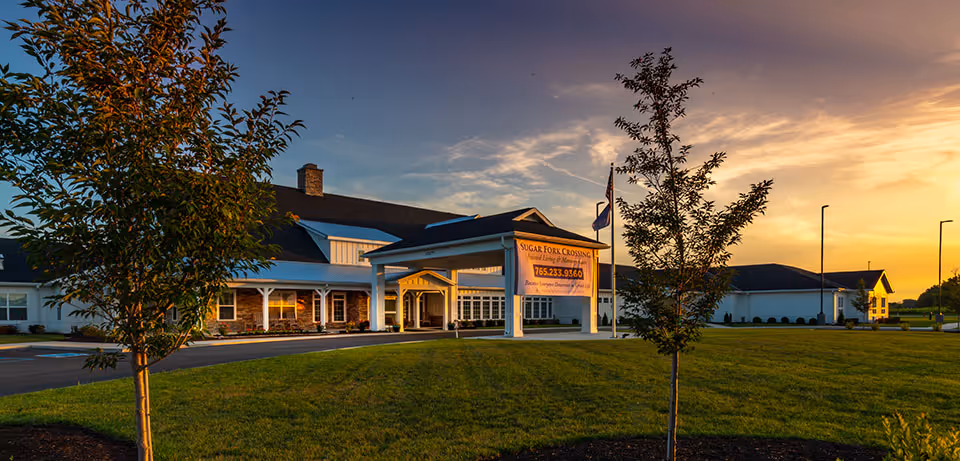 Exterior view of Sugar Fork Crossing senior living facility at sunset, showing a large building with a covered entrance, a sign with the facility's name and phone number, a well-maintained lawn, and young trees in the foreground.