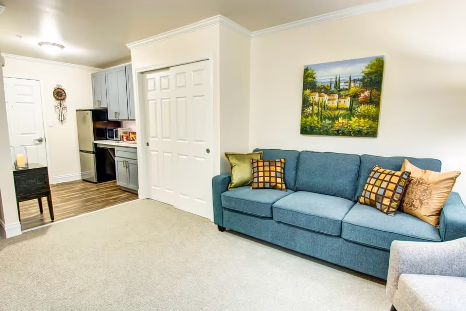 Interior view of a living area in Bridgeport Place Assisted Living featuring a blue sofa with decorative pillows, a gray armchair, a colorful landscape painting on the wall, and a small kitchen area with gray cabinets, a refrigerator, microwave, and wooden flooring near the entrance door.