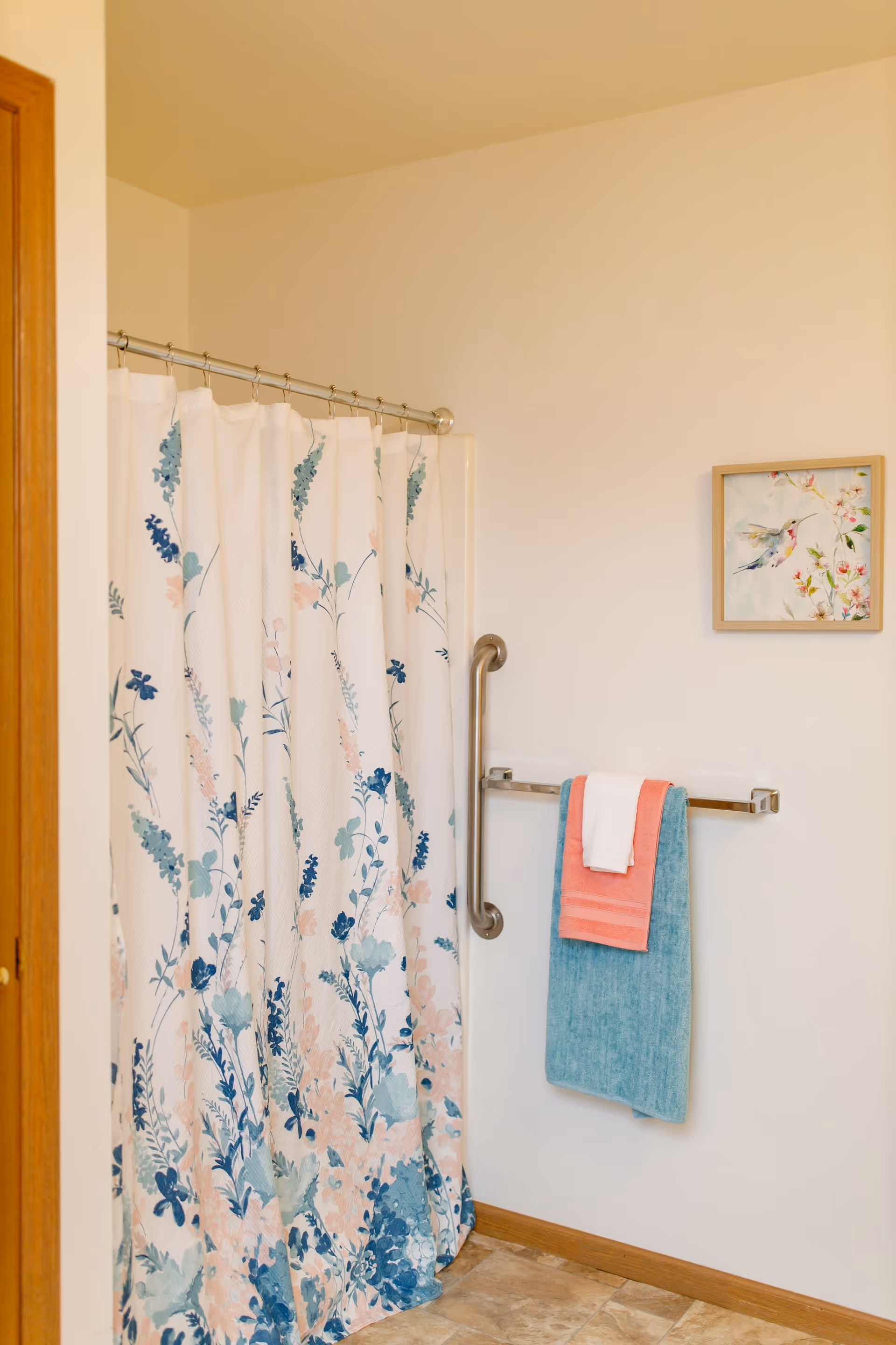 Bathroom with a floral patterned shower curtain, a metal grab bar, a towel rack holding three towels in white, coral, and teal, and a framed picture of a hummingbird on the wall.
