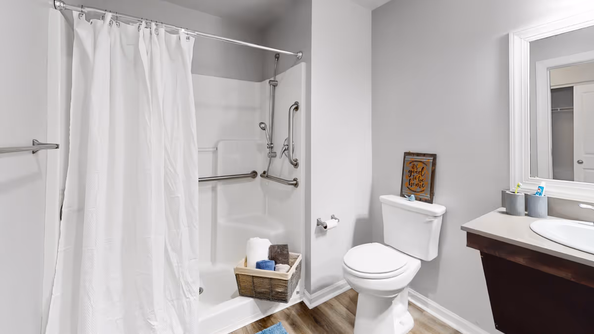 Accessible bathroom with a shower and curtain, toilet, sink vanity with mirror, grab bars, and a basket of towels.