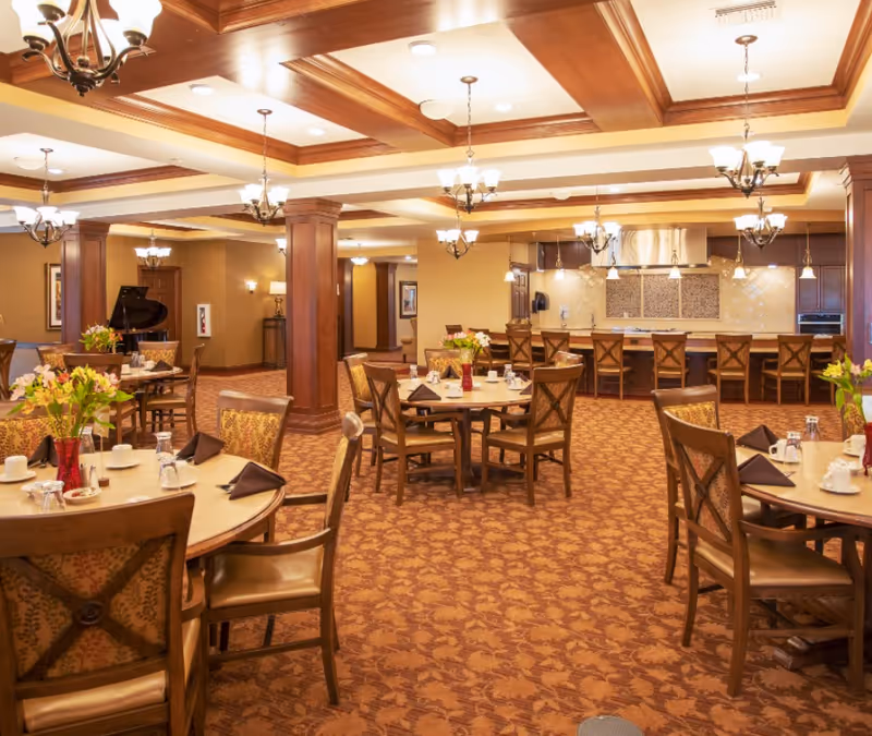 Spacious, warmly lit dining room with round tables and chairs, chandeliers, and a serving counter/kitchen in the background.