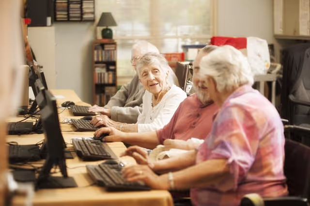 A group of elderly people sitting side by side at a long table using desktop computers in a well-lit room, engaging with each other and smiling.