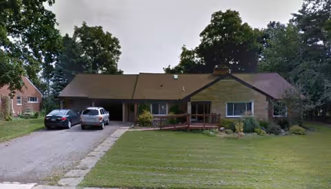 A single-story assisted living facility with a driveway, parked cars, and a well-kept lawn.