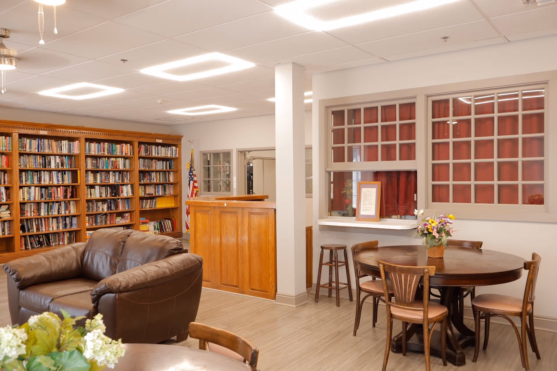A cozy senior living common area with a brown leather loveseat, a round wooden table with four chairs and a flower arrangement, a wooden reception desk, and a large bookshelf filled with books. There is an American flag in the corner and windows with red curtains behind the reception desk.