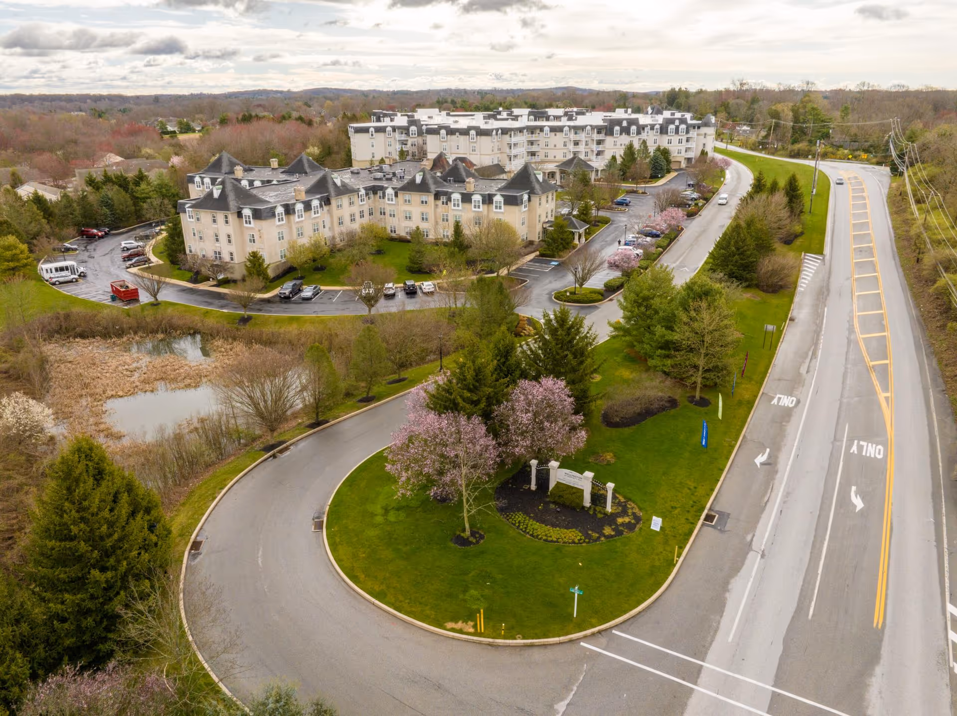 Aerial view of a large residential care building with a curved entrance drive, landscaped grounds, parking, and a nearby roadway.