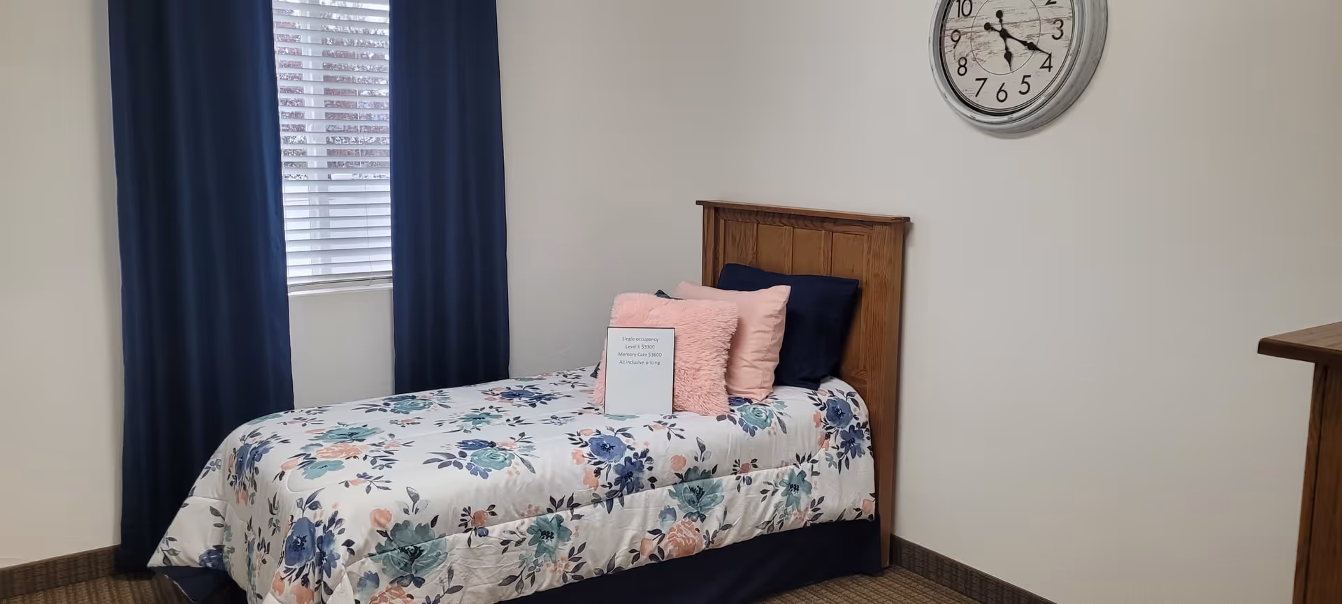 A small bedroom with a single bed featuring a floral bedspread and several pillows in shades of pink and navy blue. The bed has a wooden headboard. There is a window with white blinds and navy blue curtains on the left wall. A round clock with a white face and black numbers hangs on the plain white wall above the bed. The floor is carpeted in a neutral tone.