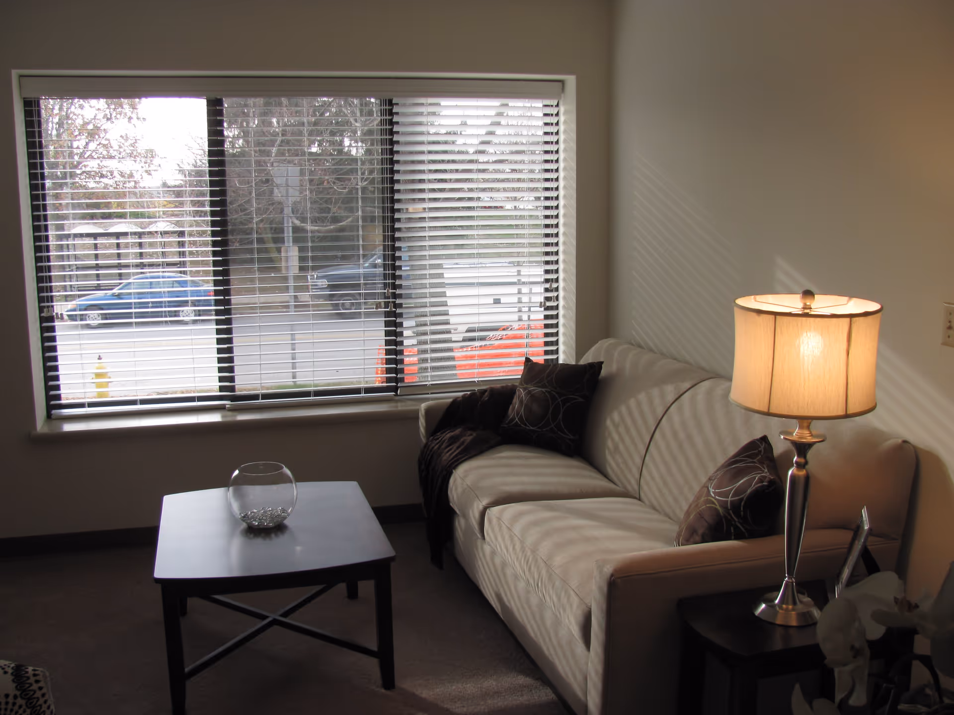 A living room with a beige sofa, side table and lamp, coffee table, and a large window with blinds showing a street outside.