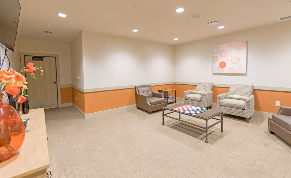 A cozy lounge area with beige carpet and walls painted white with an orange accent strip. The room features two patterned armchairs and two brown leather chairs arranged around a coffee table with a checkerboard game set on top. A small side table with board games is placed between the chairs. A flat-screen TV is mounted on the wall above a wooden cabinet with an orange vase and flowers. A piece of abstract art with circular patterns hangs on the wall.