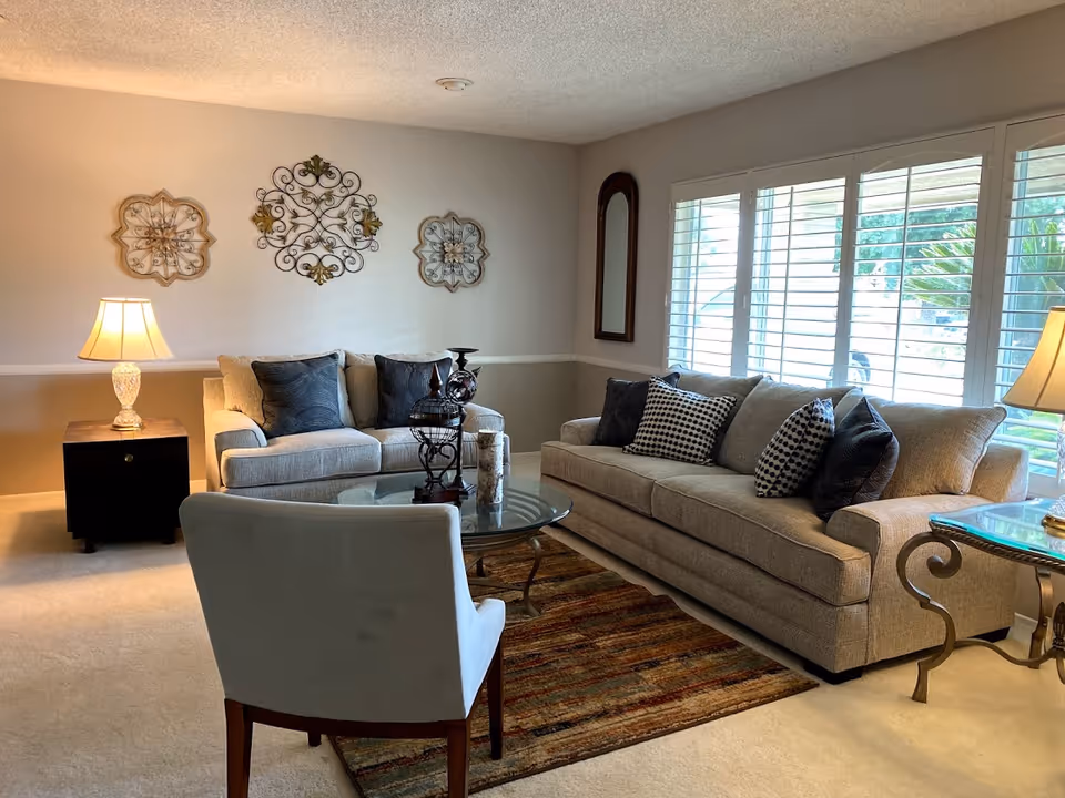 Bright living room with two gray sofas, an armchair, glass coffee table, lamps and decorative wall art by shuttered windows.