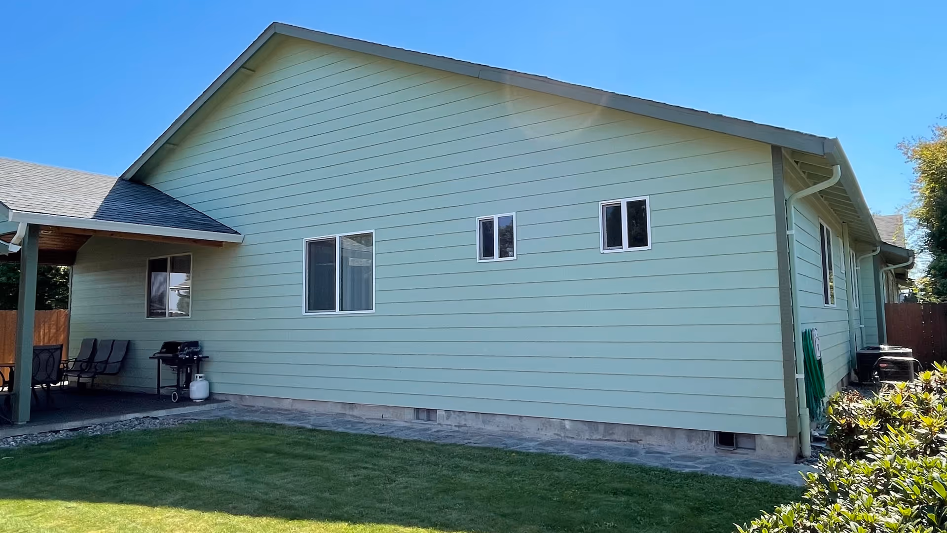Side exterior of a single-story light green house with several windows, a covered patio with chairs and a grill, and a grassy yard.