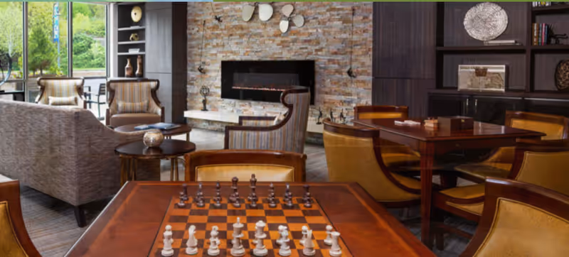 Cozy common lounge with a chessboard on a wooden table, upholstered chairs and sofas, and a fireplace set into a stone accent wall.