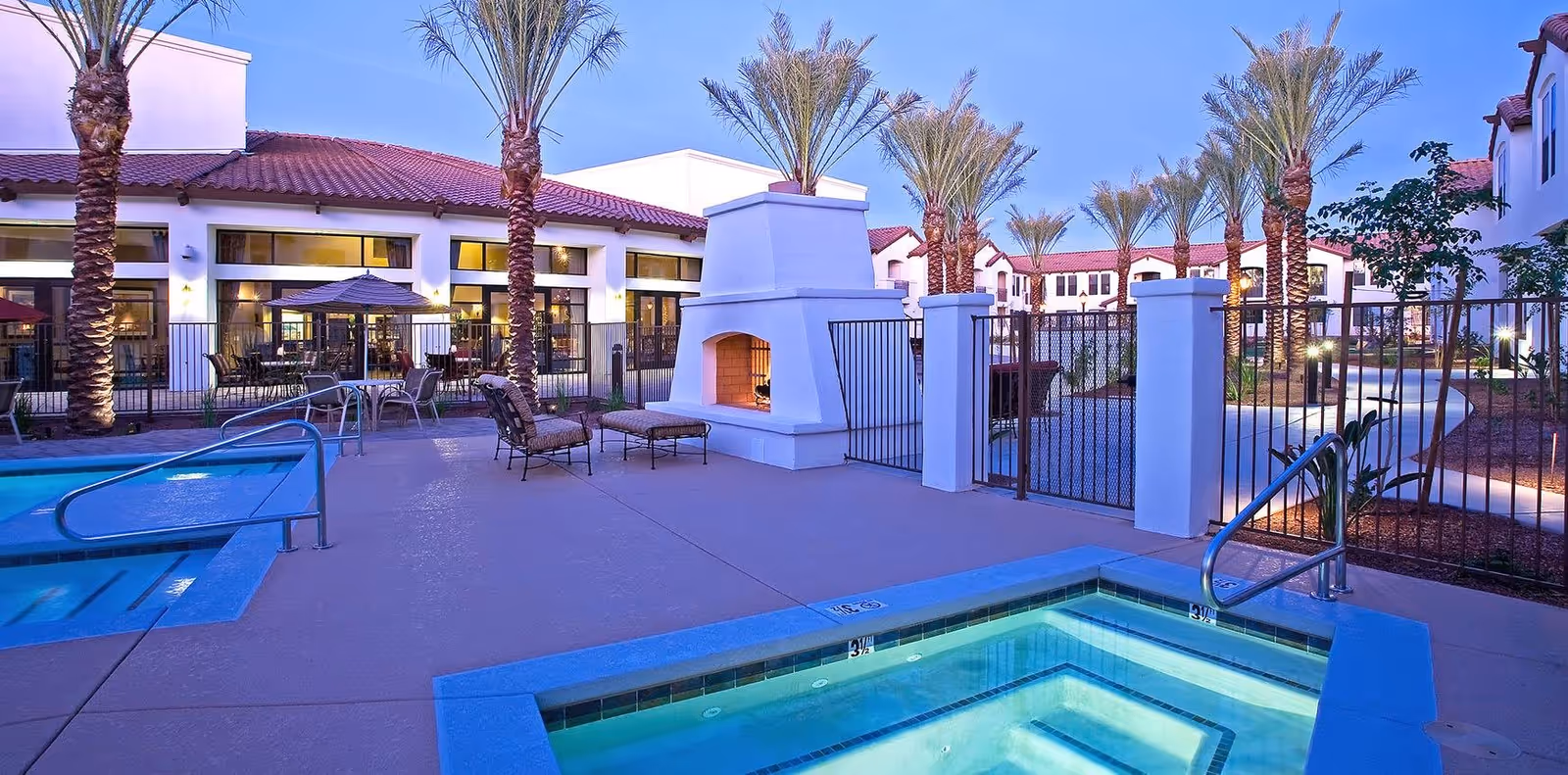 Outdoor resort-style pool and hot tub area with seating, a white fireplace, palm trees, and the facility building at dusk.