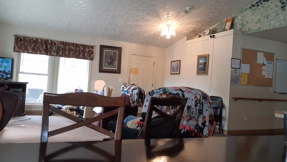 A cozy living room area with two chairs covered in colorful blankets featuring animal prints. A person is seated near large windows with patterned valances, and a television is visible on a stand to the left. The walls have framed pictures and a bulletin board with various notices. The ceiling has a light fixture, and the room has a warm, inviting atmosphere.
