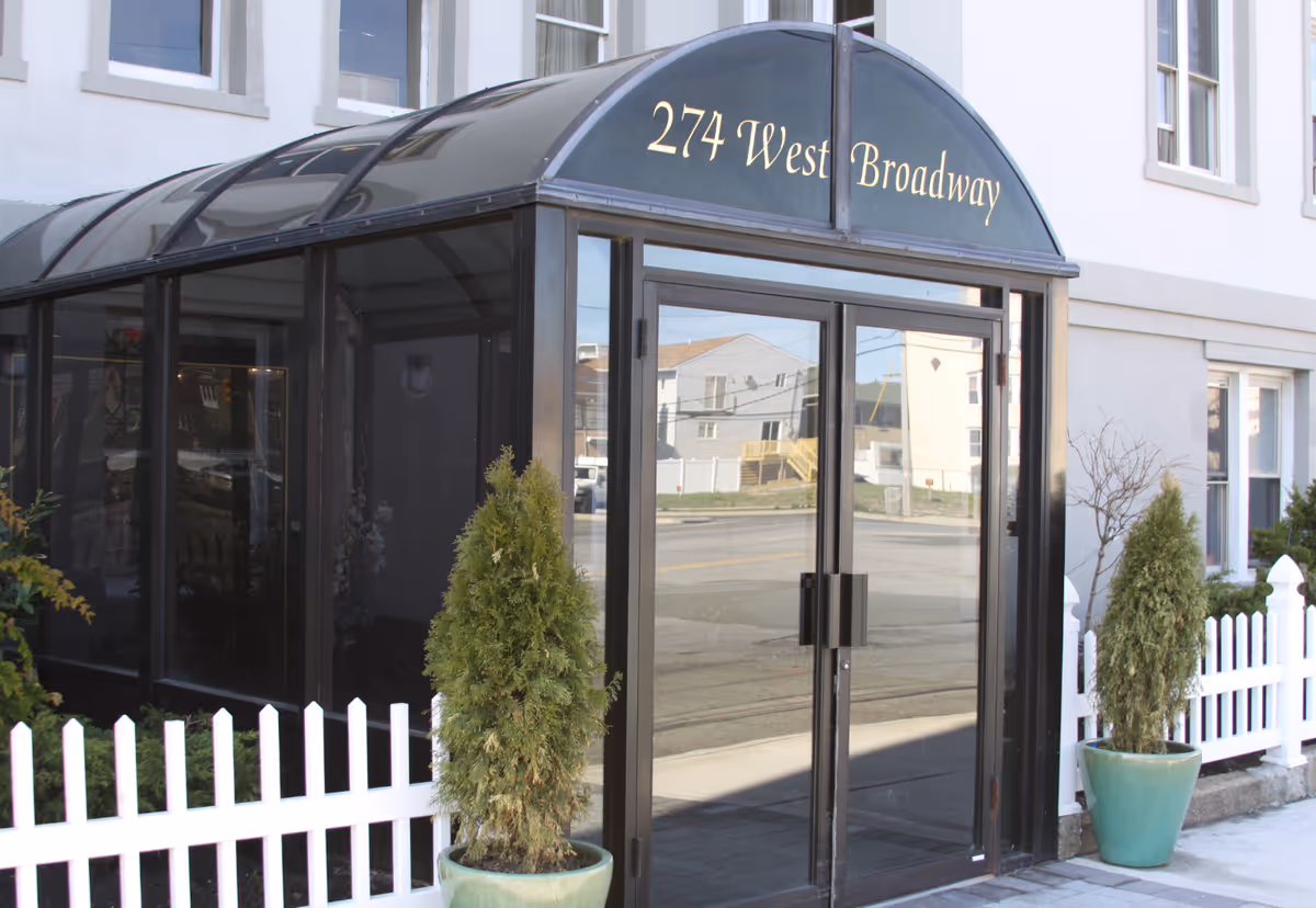 Entrance to a building with a black framed glass door and a black canopy above it displaying the address '274 West Broadway'. There are two potted evergreen plants on either side of the door and a white picket fence in front of the building.