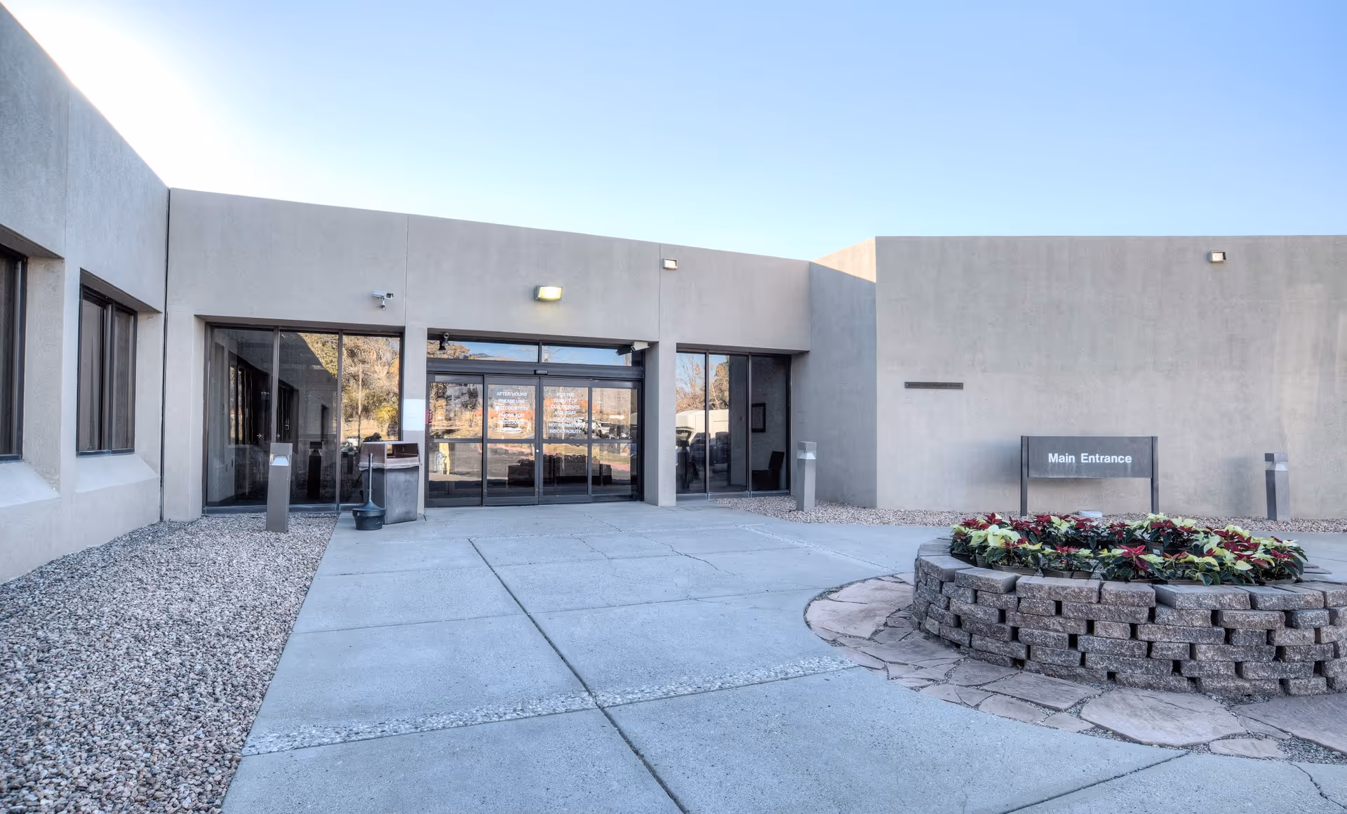 Exterior view of the main entrance to The Rehabilitation Center of Albuquerque, featuring a modern building with large glass doors, a circular flower bed with colorful plants, and a clear blue sky.