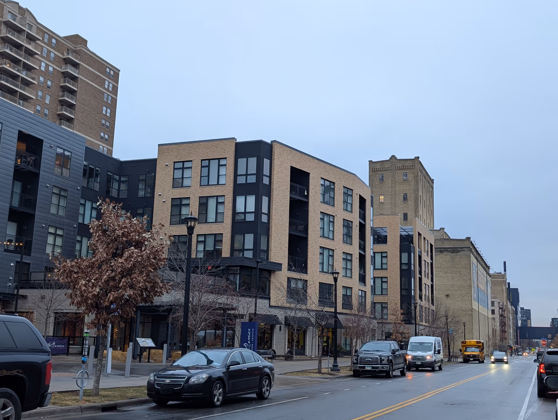 Street view of a modern multi-story residential building with beige and black exterior walls, several windows, and balconies. Cars are parked along the street in front of the building, and leafless trees line the sidewalk. The sky is overcast.