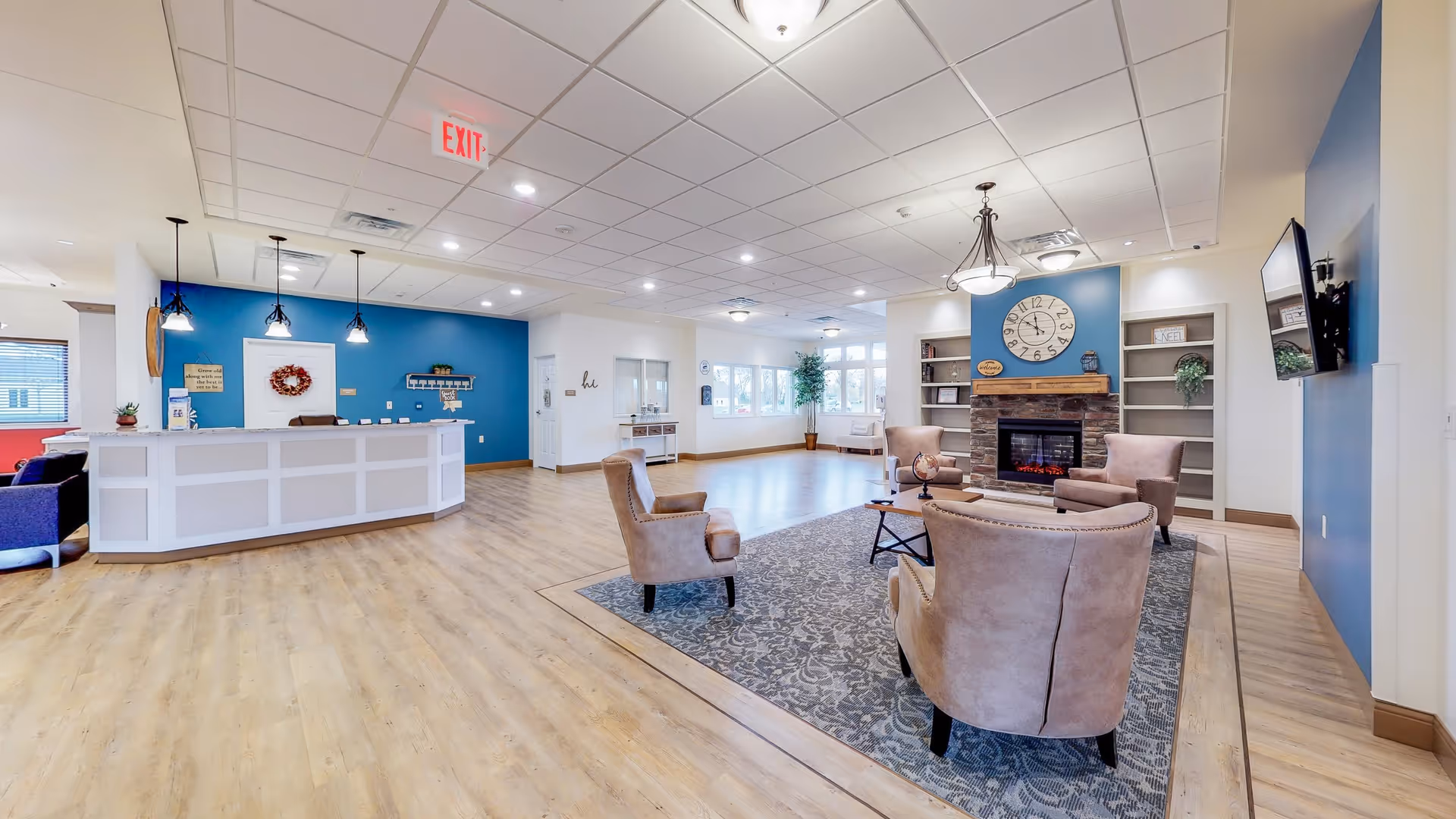 A spacious and well-lit senior living facility common area with light wood flooring and a blue accent wall behind a white reception desk. The seating area includes four beige armchairs arranged around a small coffee table on a patterned rug in front of a stone fireplace with a large clock above it. Built-in shelves flank the fireplace, and a flat-screen TV is mounted on the adjacent wall. Large windows allow natural light to fill the room.