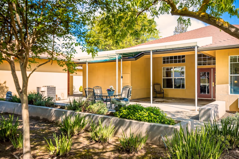 Outdoor patio area at Pacifica Senior Living Modesto featuring a covered seating area with a table and chairs, surrounded by greenery and trees under a sunny sky.