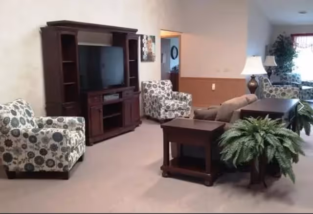 A cozy living room area with two patterned armchairs, a brown sofa, a dark wooden TV stand with a flat-screen TV, two matching dark wooden side tables, a table lamp, and a potted fern. The room has beige walls and carpeted flooring, with a doorway leading to another room.