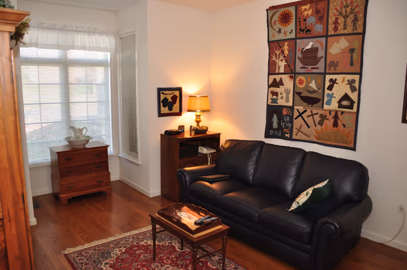 A cozy living room with a black leather couch adorned with two pillows, a small wooden coffee table with a remote control and a decorative tray, a wooden side table with a lamp and some items on it, and a wooden chest of drawers near a window with blinds and a sheer curtain. A colorful quilt with various designs hangs on the wall above the couch.