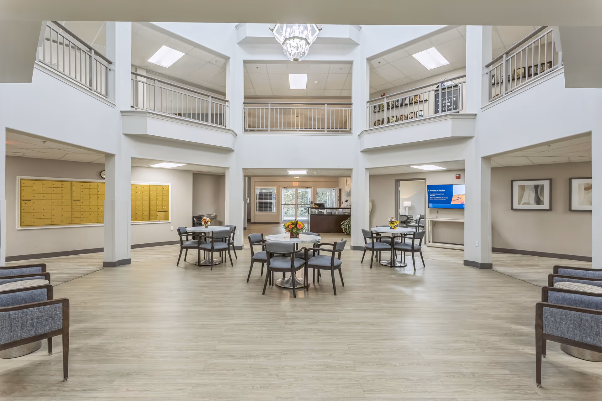 A spacious, well-lit common area in a senior living facility with multiple round tables and chairs arranged in the center. The room features a high ceiling with a chandelier, a second-floor balcony with railings, and a reception desk visible in the background. There are mailboxes on the left wall and framed artwork on the right wall.