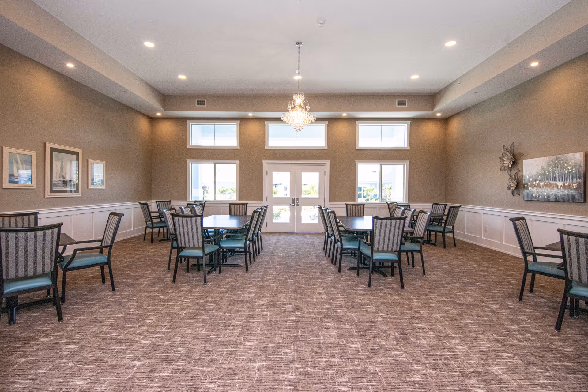 Spacious dining/activity room with multiple tables and chairs, a central chandelier, and windows on the far wall.