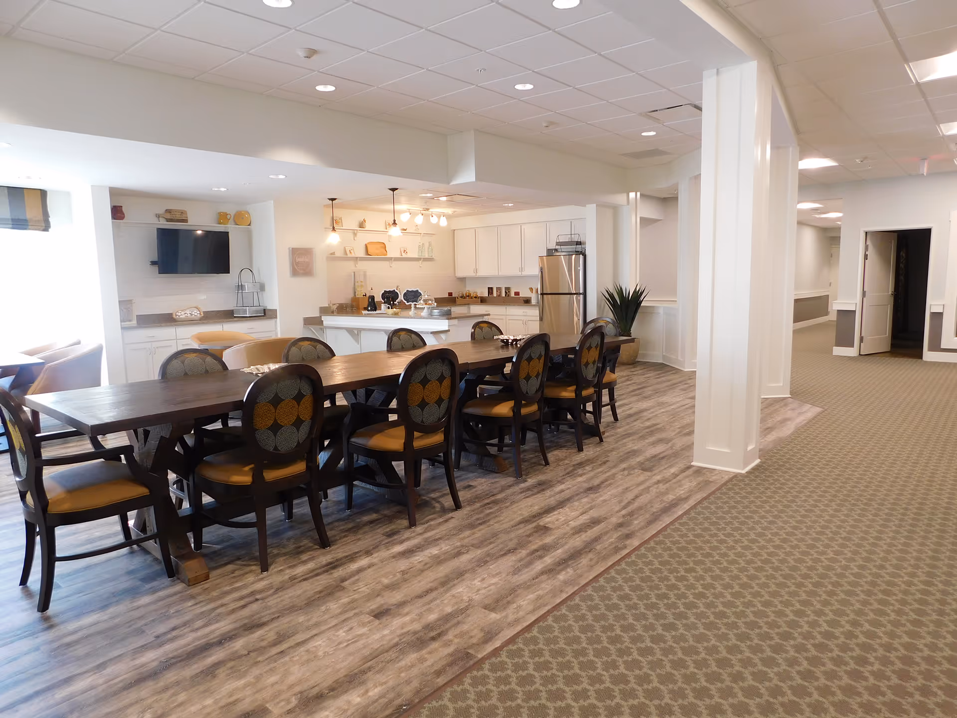 Open communal dining area with a long wooden table and chairs beside a kitchen and seating area in a senior living facility.