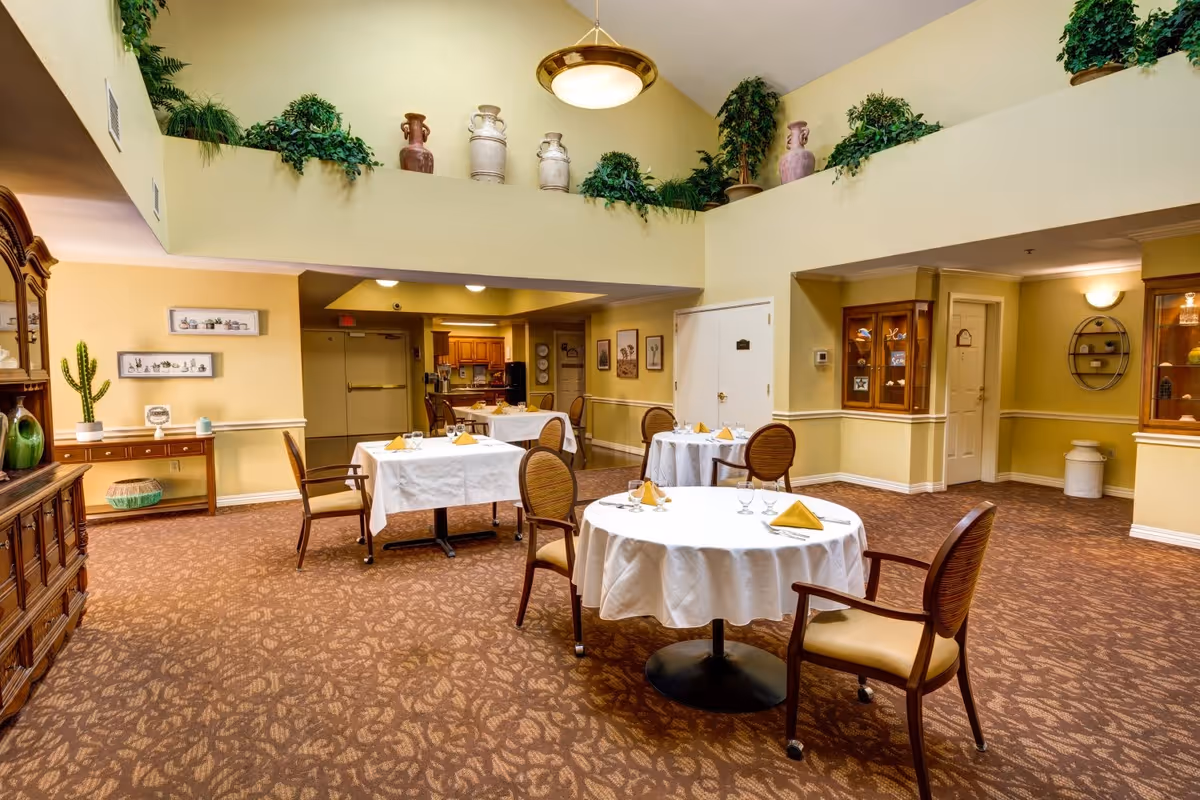 A dining room in a senior living facility with several round and square tables covered with white tablecloths, each set with yellow napkins, glasses, and silverware. The room has beige walls, carpeted floors with a leaf pattern, and a high ceiling decorated with potted plants and large ceramic vases. There is wooden furniture along the walls, framed pictures, and a warm, inviting ambiance.