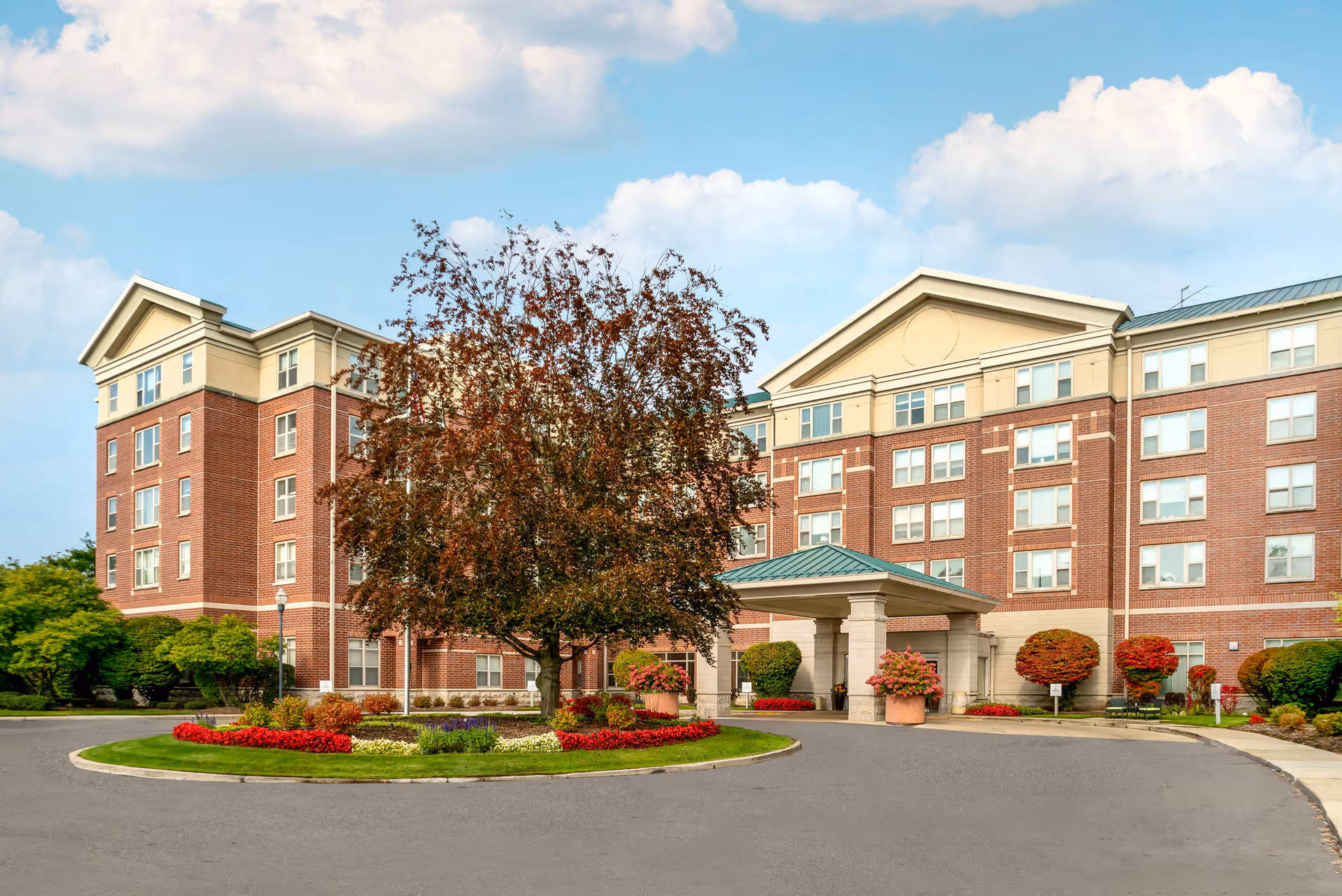 Exterior view of a multi-story senior living facility with a circular driveway, landscaped garden with a tree and flowers in the center, and a covered entrance. The building has a brick facade with numerous windows and a blue sky with some clouds in the background.