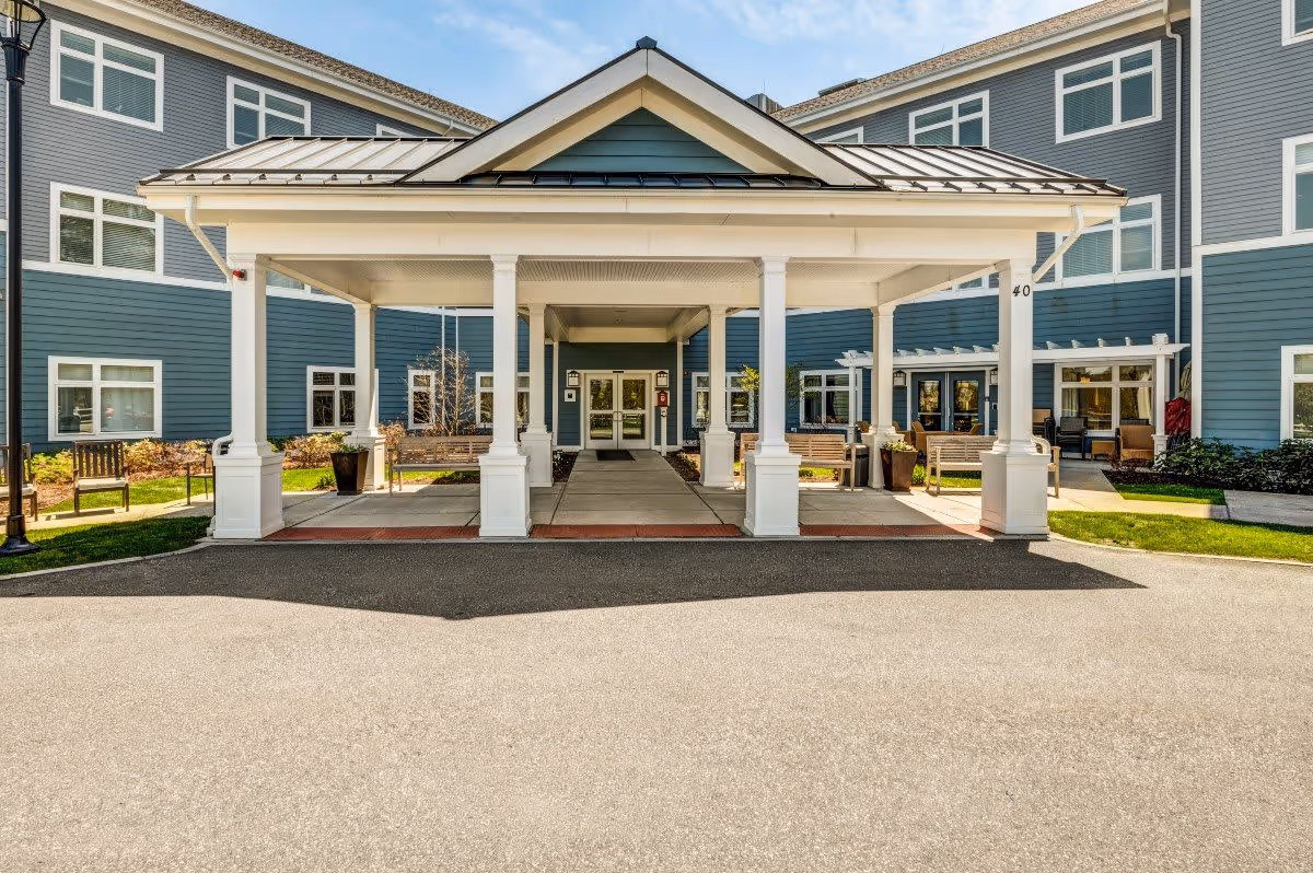 Front entrance of a senior living facility with a covered drop-off area supported by white columns. The building exterior is blue with white trim and multiple windows. There are benches and potted plants near the entrance, and a paved driveway in front.
