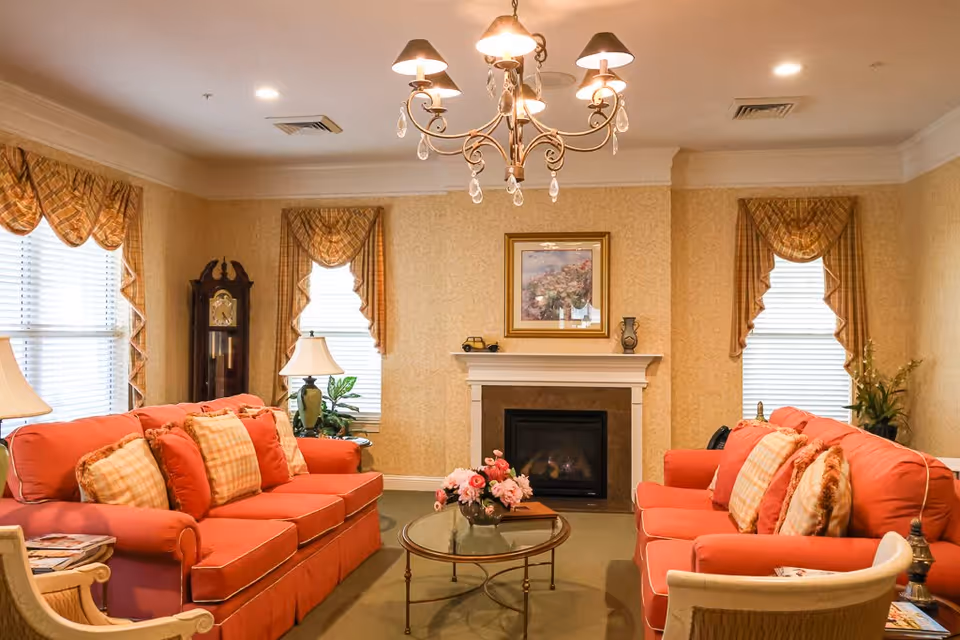 A cozy living room with two coral-colored sofas facing each other, each adorned with patterned cushions. Between them is a round glass coffee table with a flower arrangement. The room features a fireplace with a framed floral painting above it, two windows with patterned valances, a grandfather clock, table lamps, and a chandelier hanging from the ceiling.