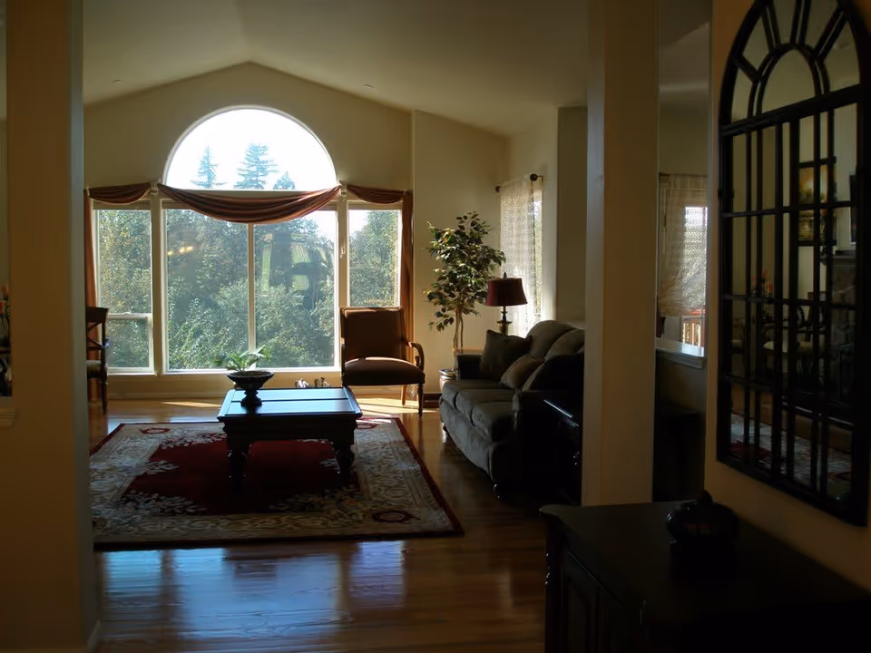 A cozy living room with a large arched window letting in natural light. The room features a sofa, an armchair, a coffee table on a patterned rug, a floor lamp, and a potted plant. There is a decorative mirror on the right wall reflecting part of the room.