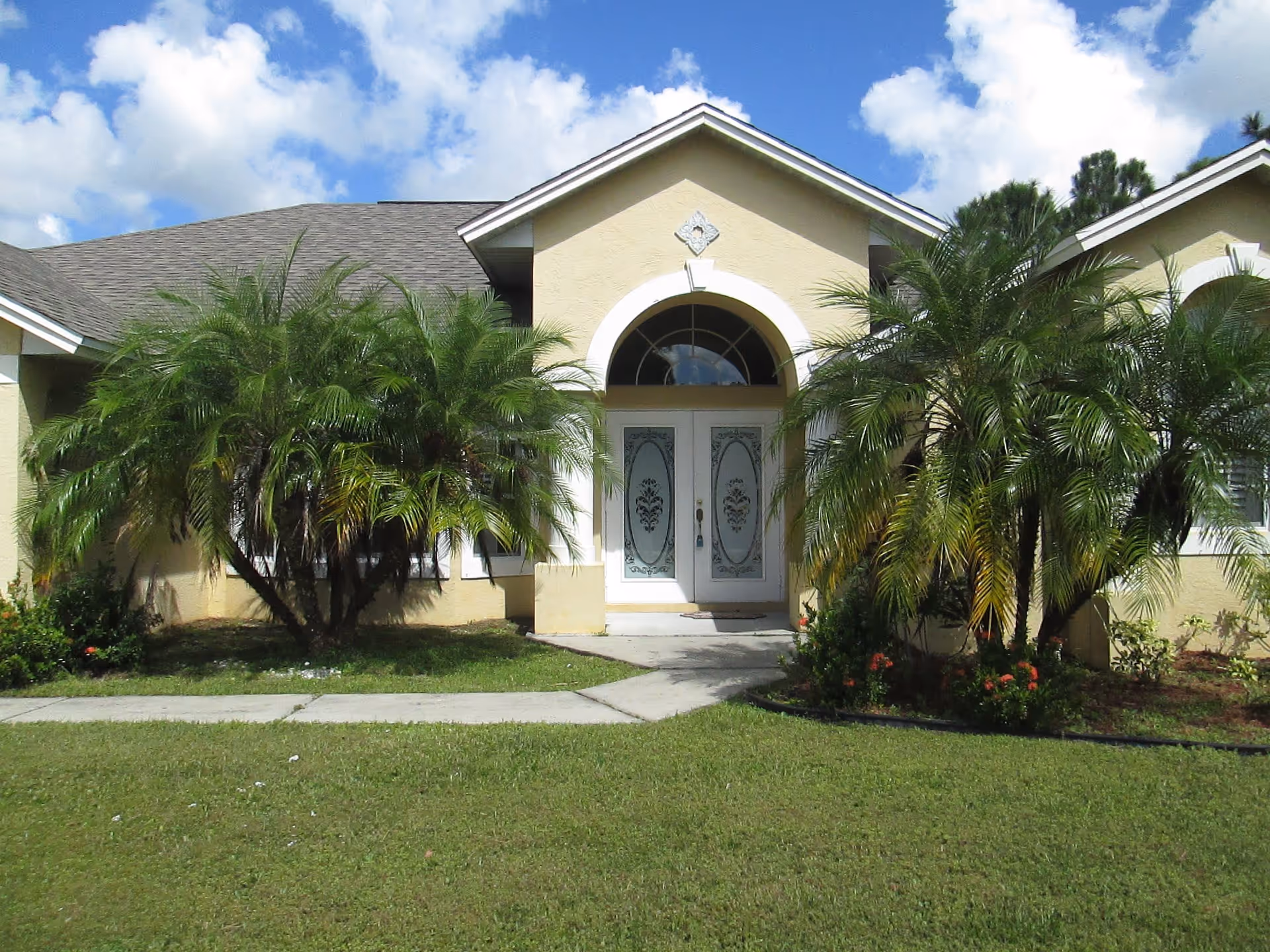 Front exterior view of a single-story building with a beige facade, a peaked roof, and decorative glass double doors. The entrance is flanked by two large palm trees and surrounded by a well-maintained lawn and small garden beds. The sky is partly cloudy with blue patches.