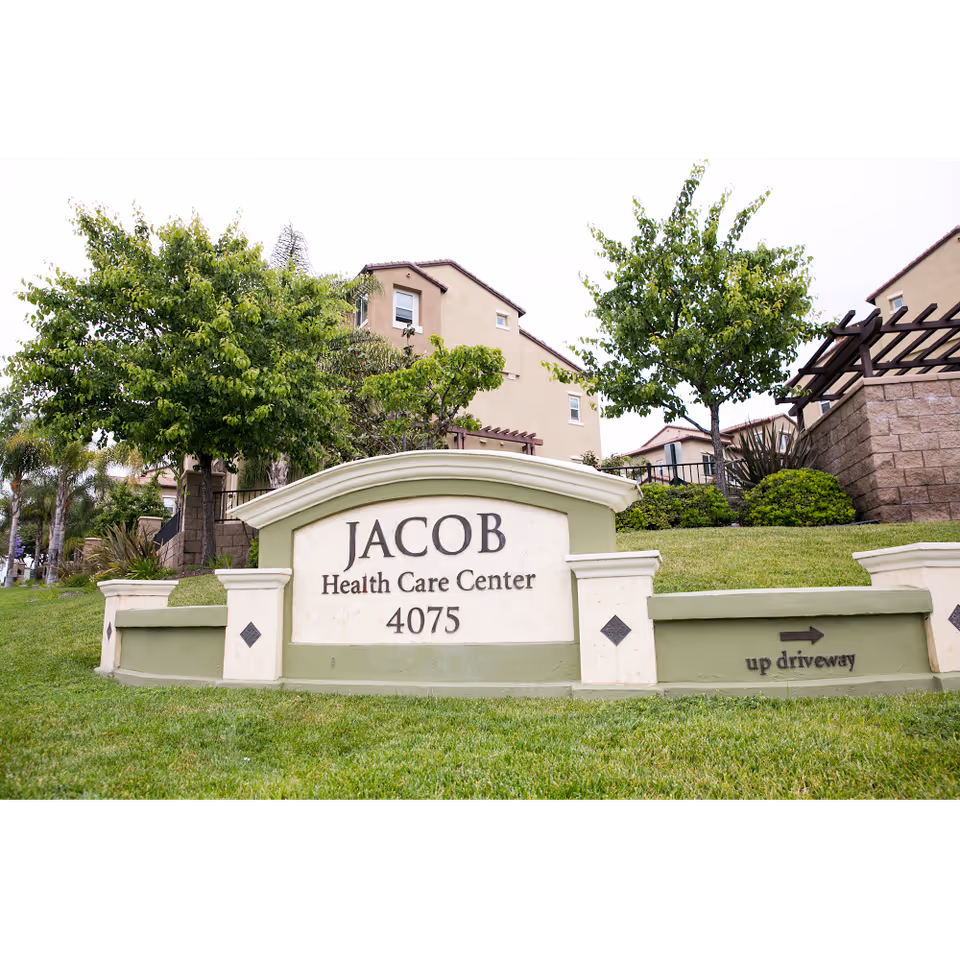 Outdoor view of the entrance sign for Jacob Health Care Center with the address 4075. The sign is surrounded by green grass, trees, and shrubs, with residential-style buildings in the background.