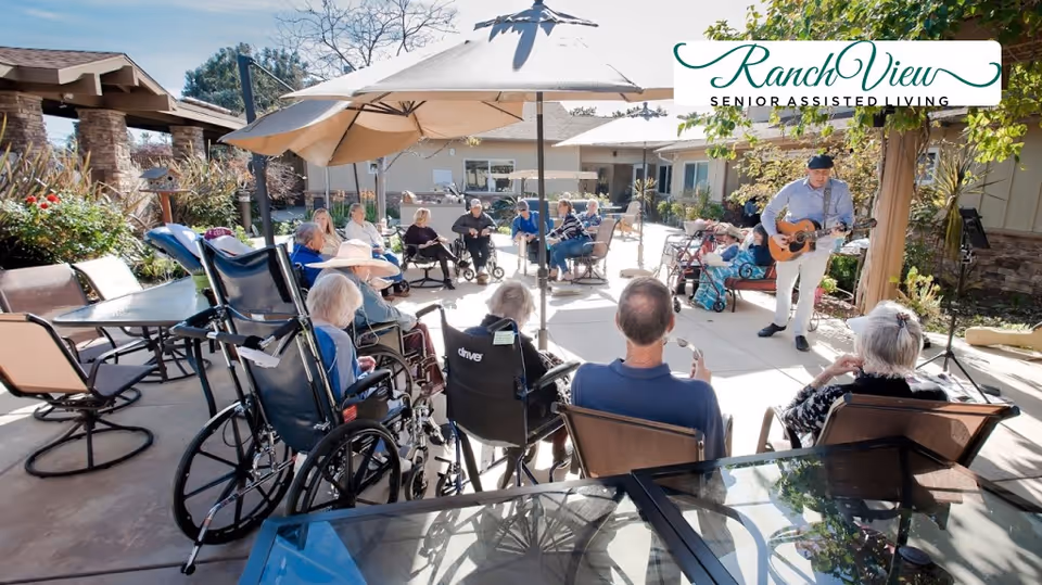 A group of elderly residents sitting outdoors in a courtyard area of a senior assisted living facility, some in wheelchairs and others in chairs, watching a man playing a guitar under a large patio umbrella on a sunny day.